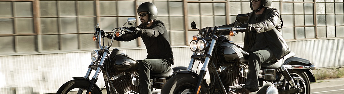 Two men in all black protective gear riding black Harley® motorcycles near an industrial warehouse. 